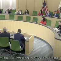 A government committee meets in a large, modern hearing room; people are seated around a curved desk. The screen displays “AB 762 Irwin: Vape ban on Disposable, Battery-Embedded Vapor Inhalation Device: Prohibition.”. CA NORML