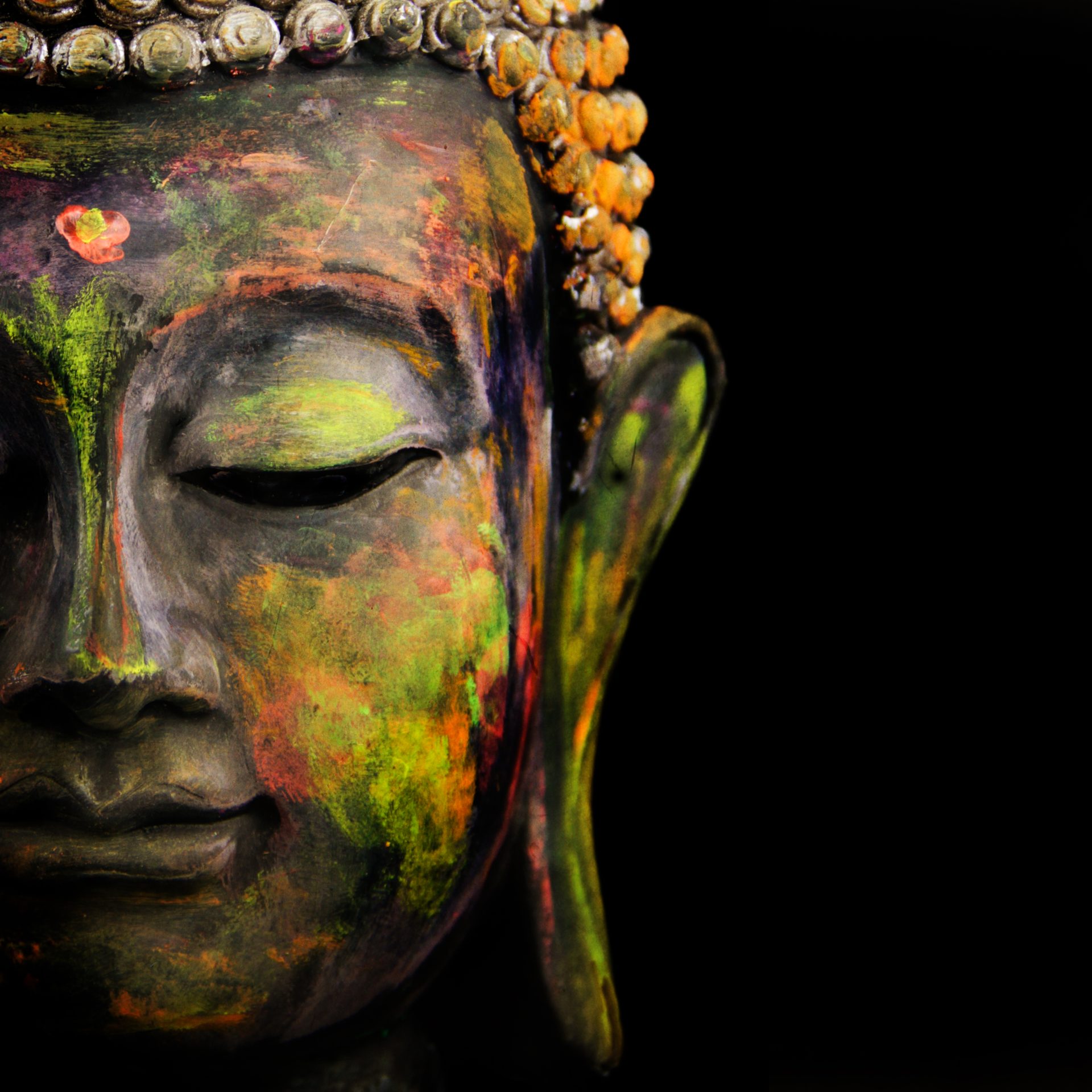 Close-up of a colorful, painted Buddha statue’s face with closed eyes, adorned with decorative beads on the crown, against a black background. Only the left half of the face is visible. CA Norml