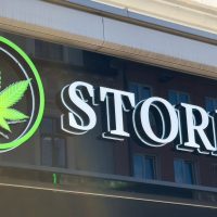 Storefront sign with the word "STORE" in bold white letters and two green cannabis leaf symbols in circles on either side, highlighting a licensed cannabis business above a window reflecting nearby buildings. Ca NORML