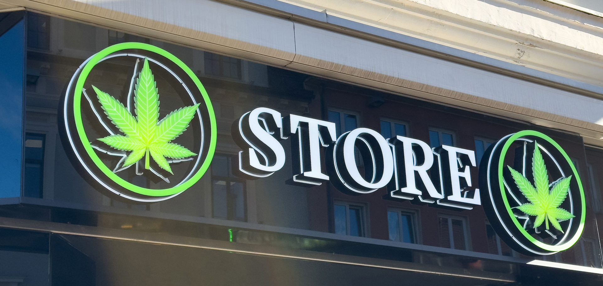 Storefront sign with the word "STORE" in bold white letters and two green cannabis leaf symbols in circles on either side, highlighting a licensed cannabis business above a window reflecting nearby buildings. Ca NORML