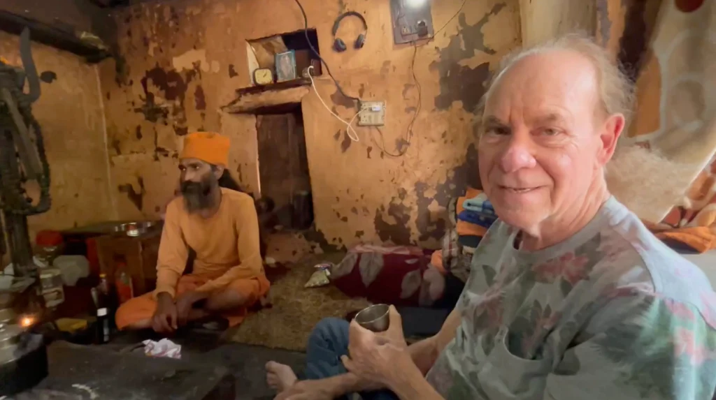 An older man holding a metal cup sits on the floor of a rustic, weathered room next to a man in orange robes and a turban. The room has peeling paint, shelves, and various items scattered around. Ca NORML