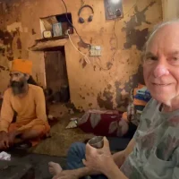 Ed Rosenthal holding a metal cup sits on the floor of a rustic, weathered room next to a man in orange robes and a turban. The room has peeling paint, shelves, and various items scattered around. Ca NORML