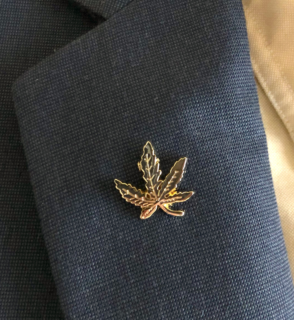 A gold pin in the shape of a cannabis leaf, inspired by Cal NORML advocacy, is attached to the lapel of a dark blue textured suit jacket. Ca NORML