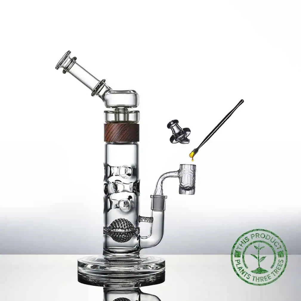 A clear glass water pipe with a wooden accent, featuring a bowl, carb cap, and dab tool with concentrate. A green stamp in the corner reads, "This product plants three trees. Ca NORML