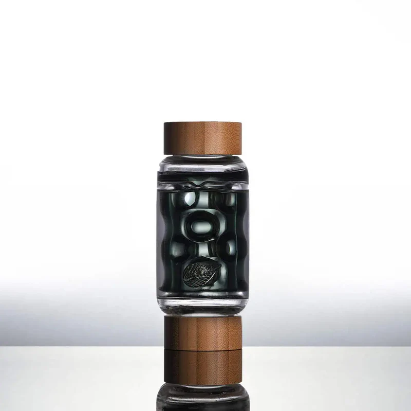 A clear glass bottle with rounded edges and a wooden cap is filled with dark liquid and several silver metallic spheres. The bottle stands upright on a reflective surface against a white gradient background. Ca NORML