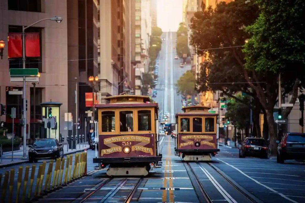 Two classic San Francisco cable cars travel up a steep city street at sunset, surrounded by tall buildings and trees, with cars parked along the road, capturing the timeless charm of San Francisco’s iconic cable cars. Ca NORML